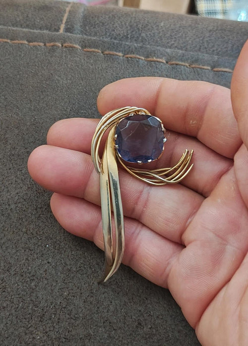 Gold-tone abstract brooch pin with blue gemstone in swirl frame, held in hand