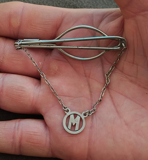 Silver tone monogram M bar clip with curved wire frame and chain pendant.