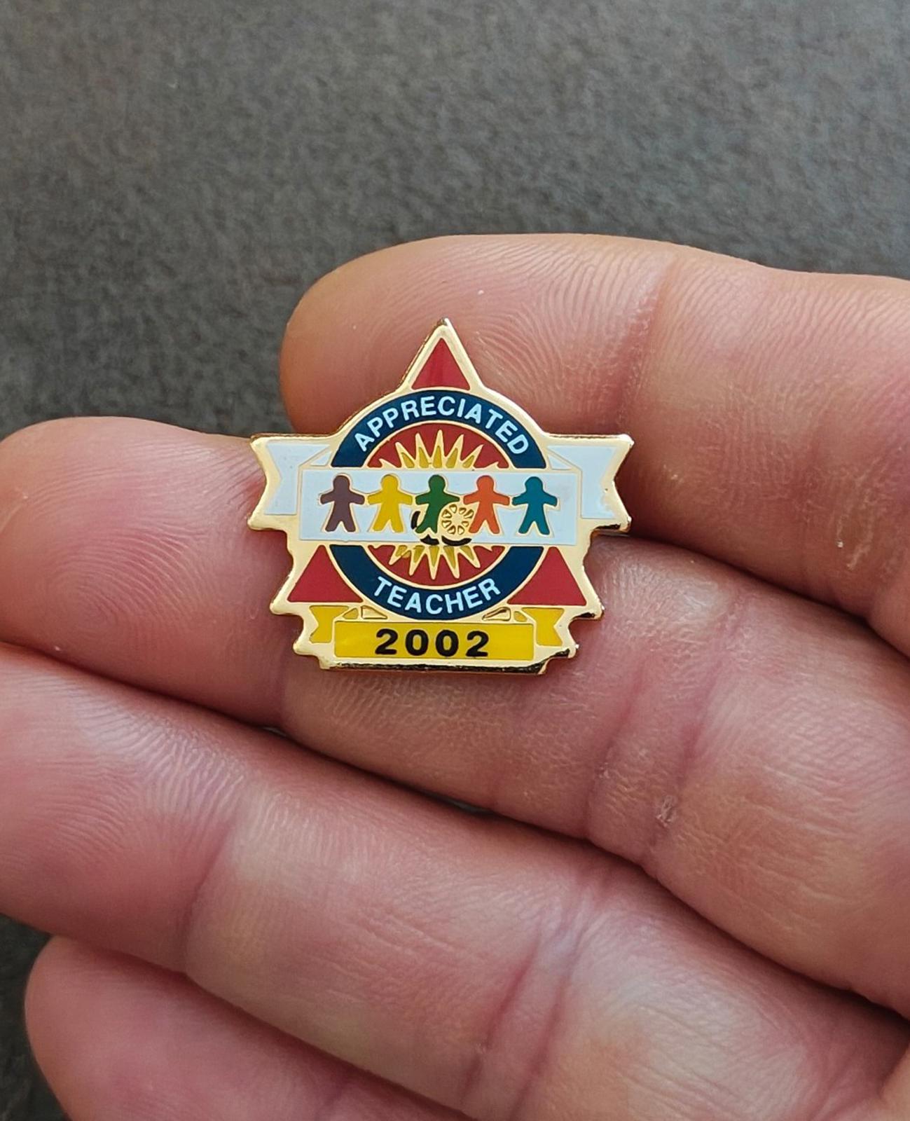 Gold-tone teacher enamel pin held in hand, blue “Appreciated” banner and “2002”