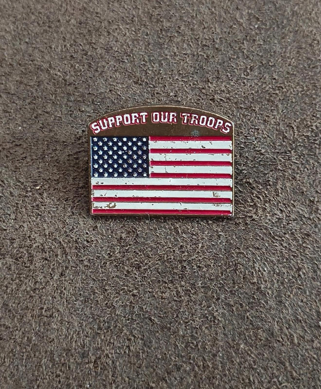 Gold-tone enamel pin on fabric with “Support Our Troops” text and U.S. flag design