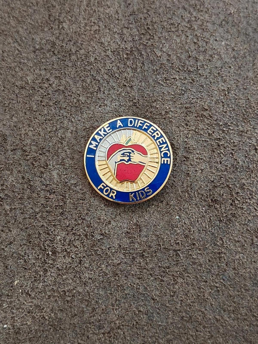 Round gold-tone enamel pin on surface: blue ring text and central apple/hand graphic.