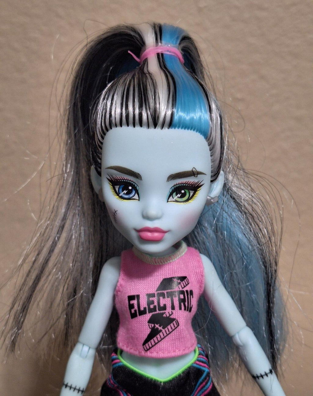 Frankie doll close-up with blue-and-black hair and pink “Electric” graphic sleeveless top.