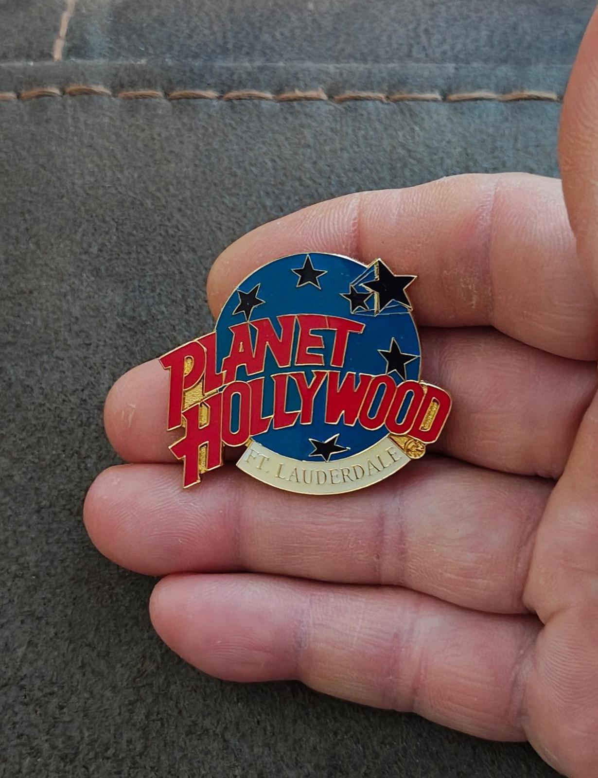 Close-up: blue enamel lapel pin with red Planet Hollywood lettering and stars, gold edge.