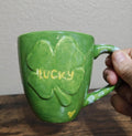 Green ceramic mug with clover-heart design and “Lucky” text, held by the handle