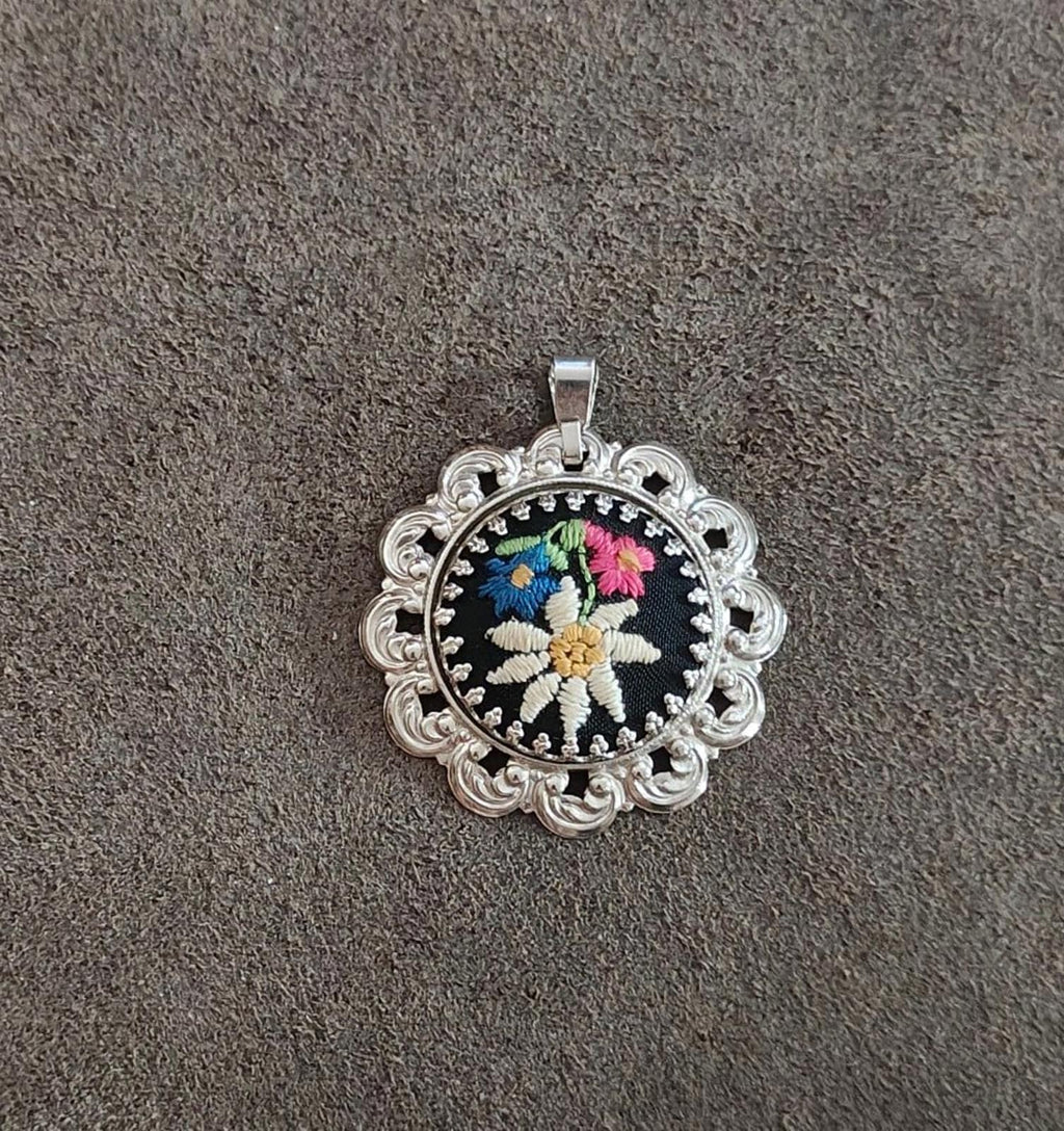 Silver filigree round pendant with black background and multicolor stitched flower.