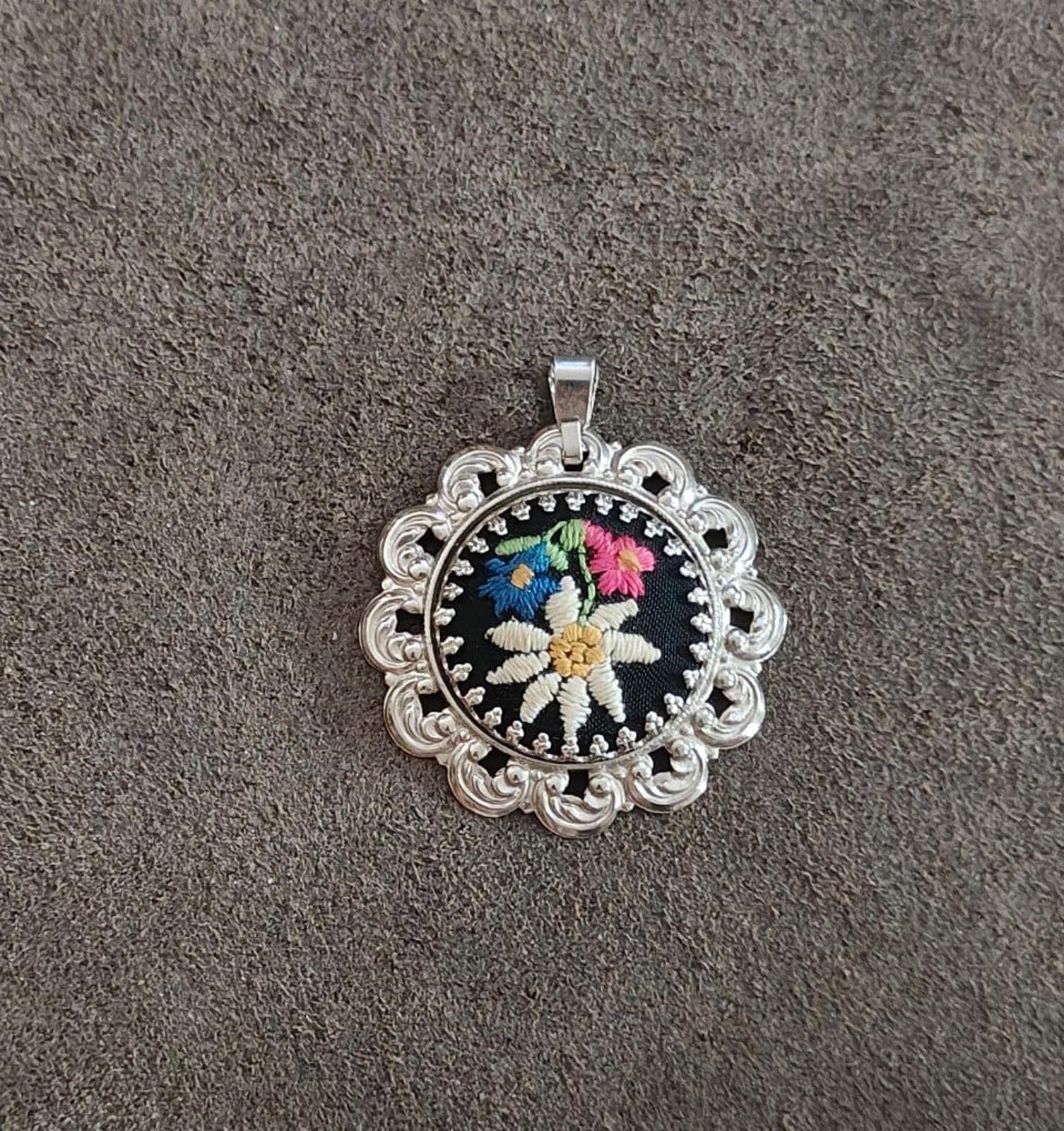 Silver filigree round pendant with black background and multicolor stitched flower.