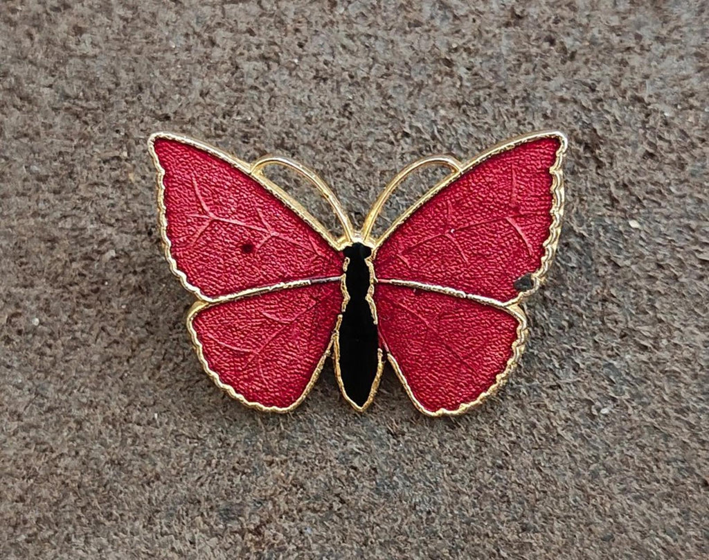 Red butterfly enamel brooch with gold-tone wings and black body, flat on textured surface.