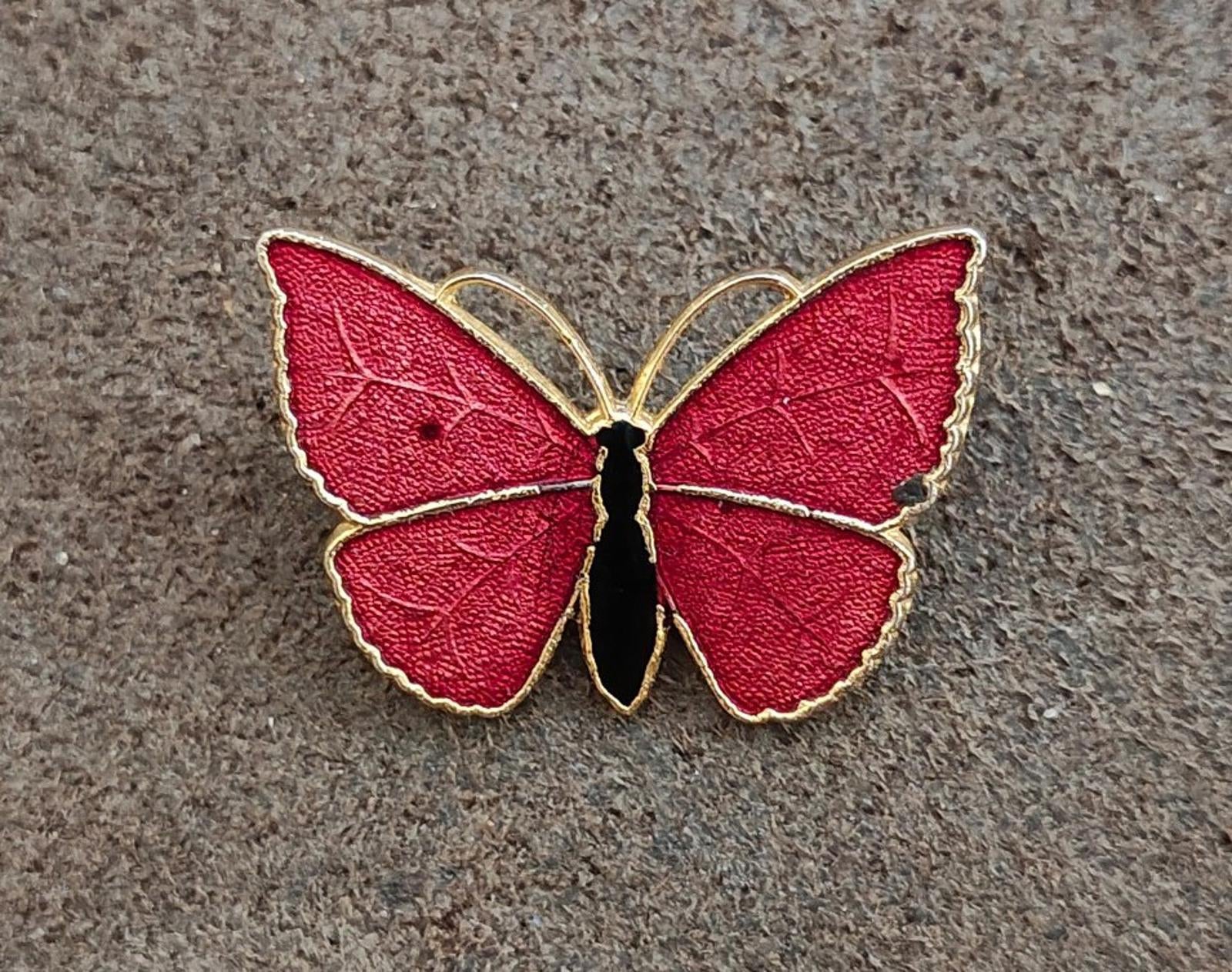 Red butterfly enamel brooch with gold-tone wings and black body, flat on textured surface.