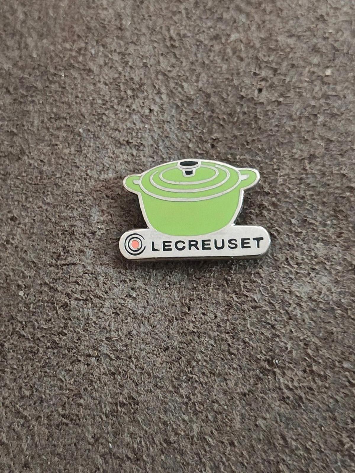 Le Creuset green mini cocotte enamel pin lying on gray fabric surface