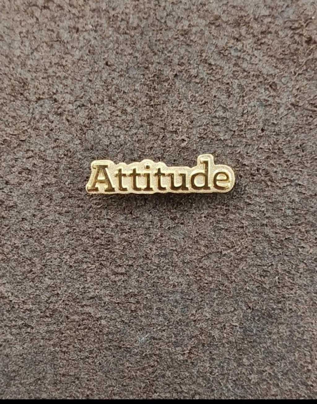 Gold tone lapel pin with raised "Attitude" lettering on gray fabric