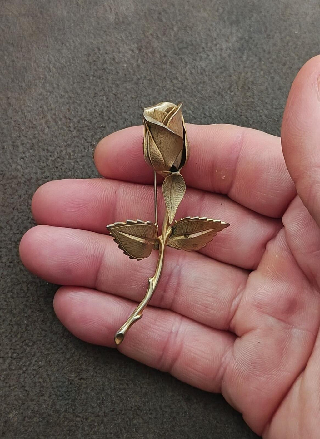 Gold tone rose brooch with central flower and curved pin stem, held in fingers