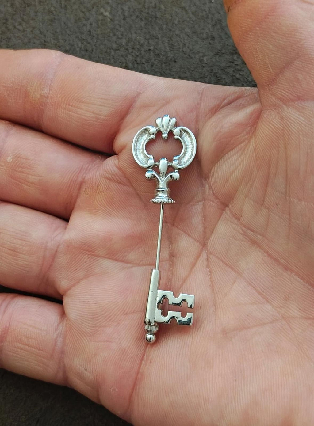 Silver fleur-de-lis stick pin with skeleton key charm, viewed in hand against skin.