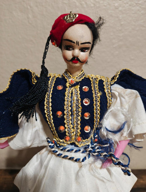 Greek folk male doll close-up: red headpiece, painted face, navy vest with gold trim