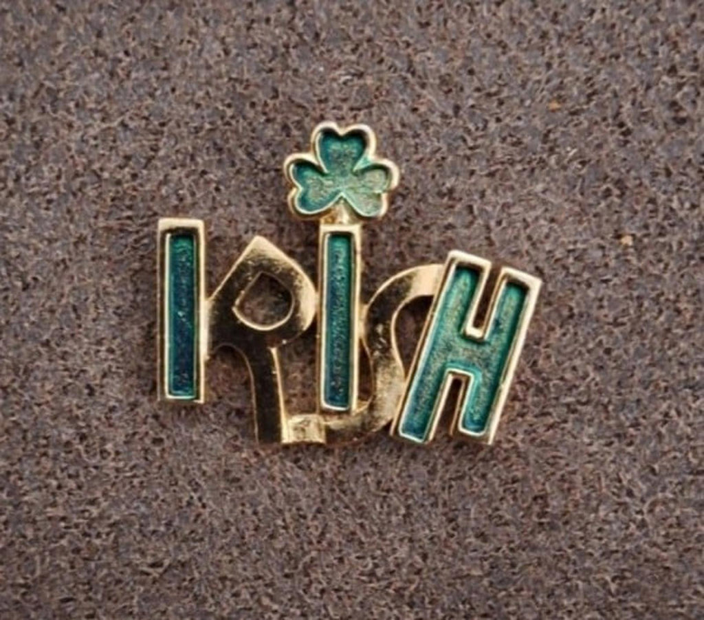 Gold-tone enamel Irish pin with green clover shamrock, angled view on textured surface.