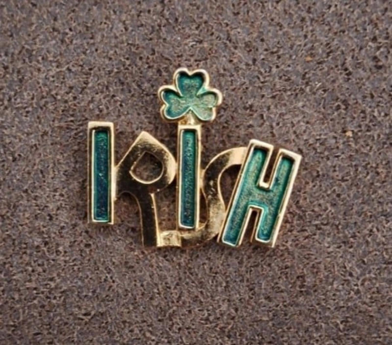 Gold-tone enamel Irish pin with green clover shamrock, angled view on textured surface.