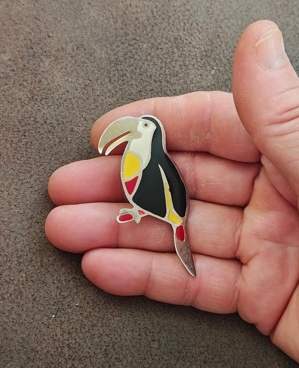 Toucan bird enamel brooch in hand, angled view; black body with yellow and red accents.
