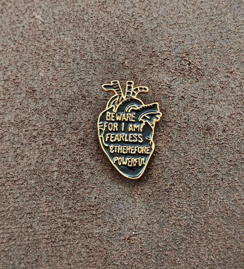 Gold-tone anatomical heart enamel pin with dark blue base and warning text on gray surface