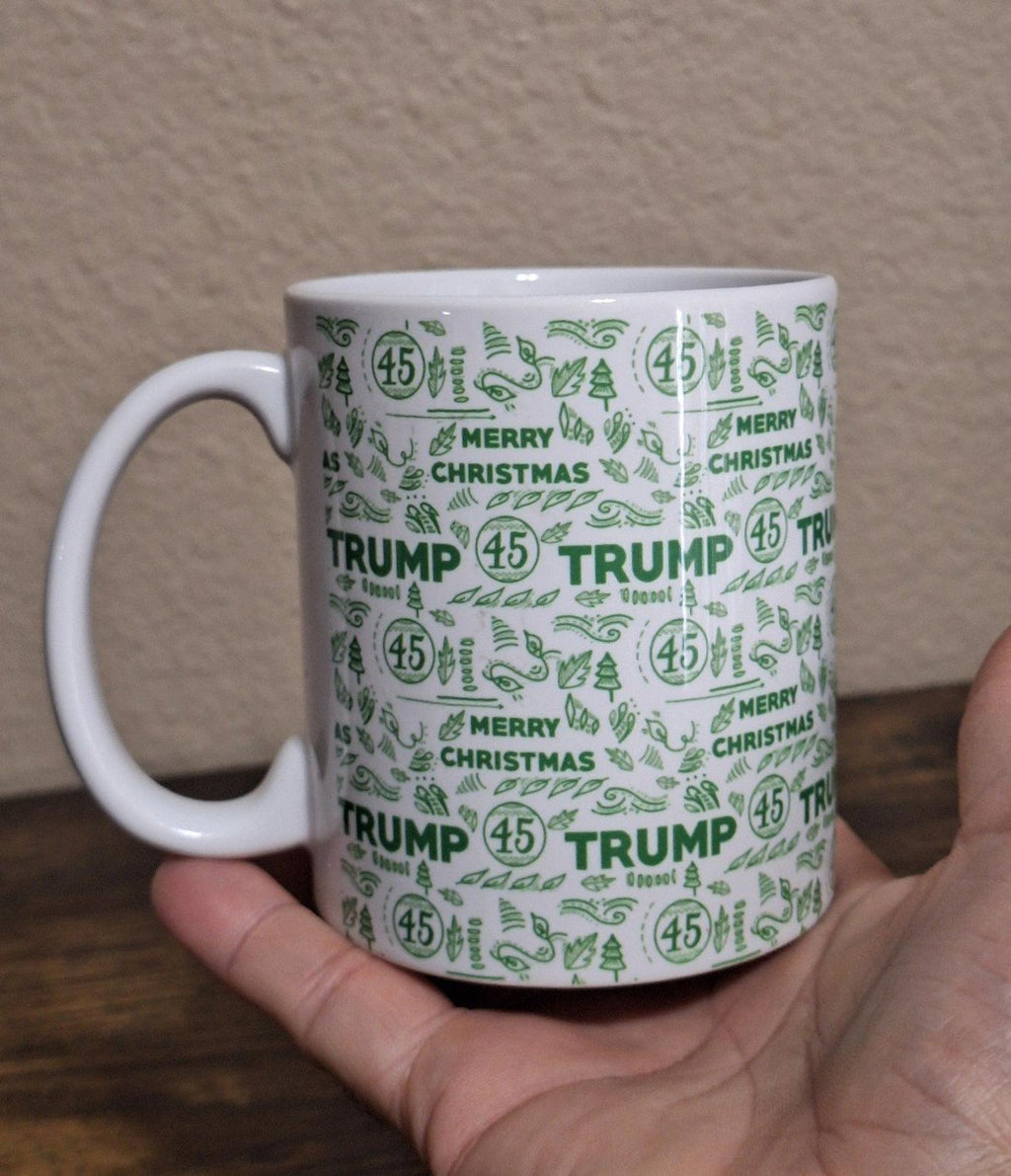 White ceramic mug with green “TRUMP” and “MERRY CHRISTMAS” repeating pattern on body.