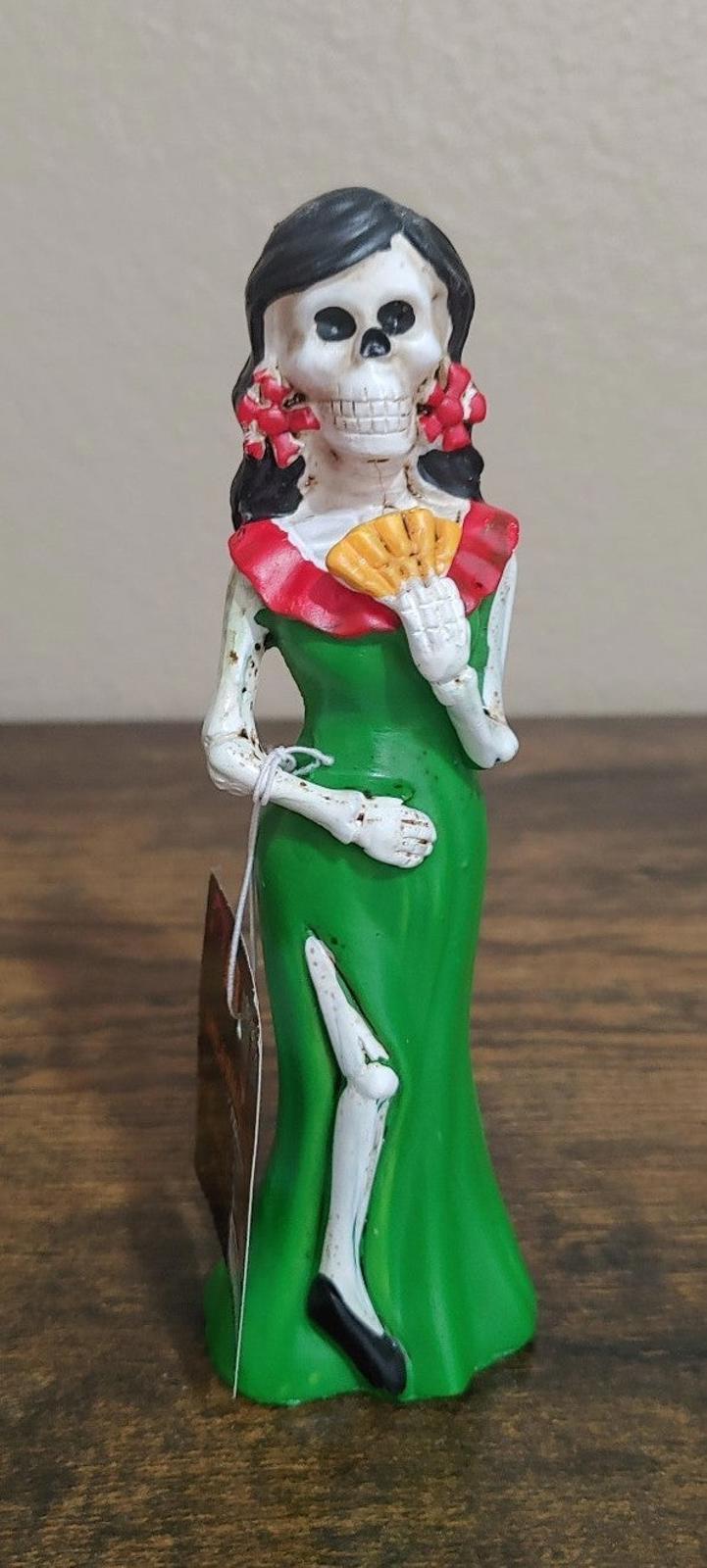 Green Catrina figurine with black hair and white skull face, holding folded food