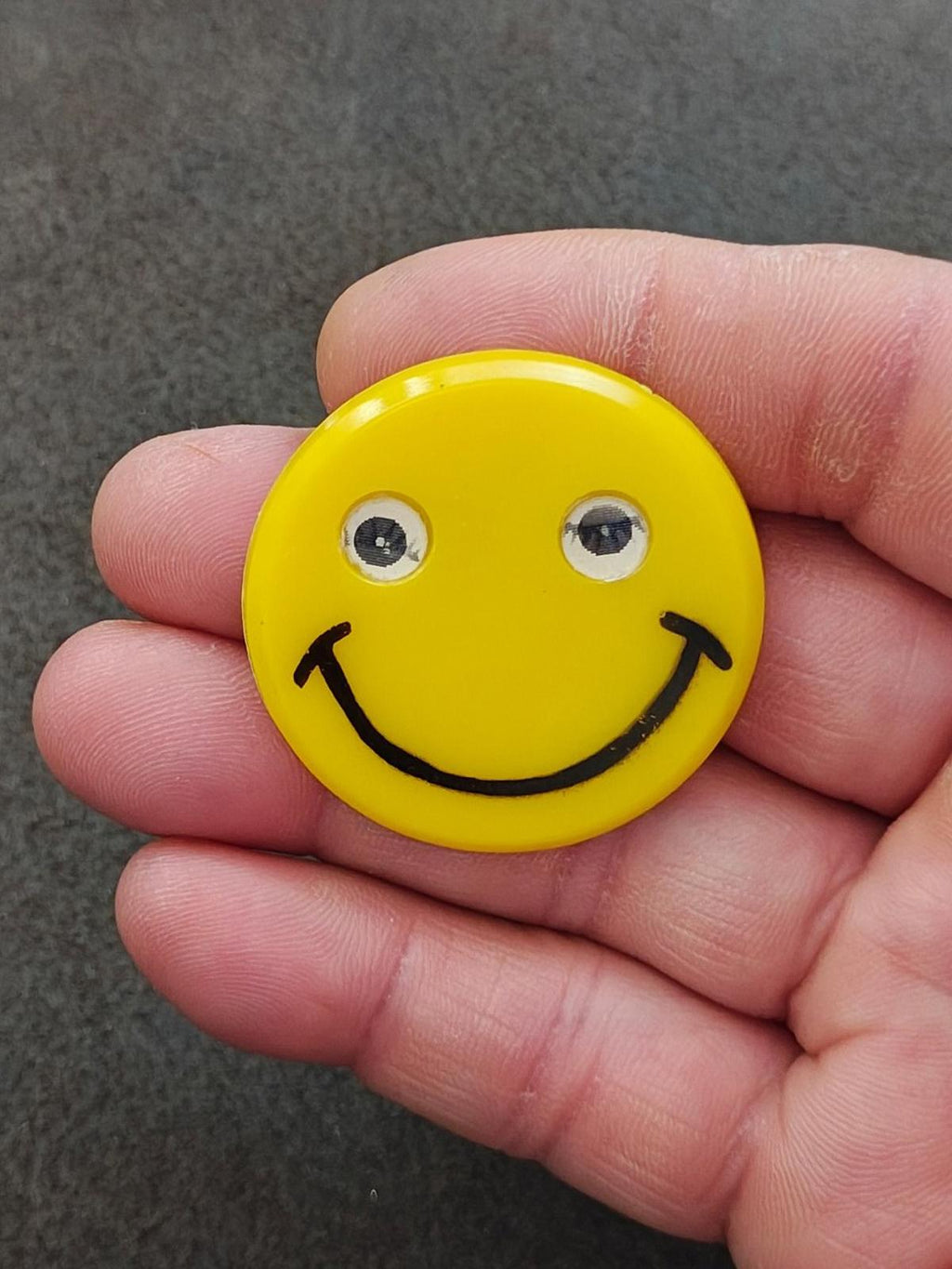 Yellow smiley face pin held in hand; black smile and mirrored eye details
