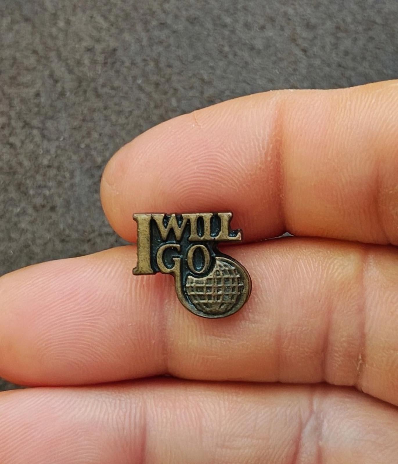 Gold tone "I WILL GO" ministry pin with globe-style emblem, held between fingers