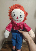 Handmade rag doll with orange yarn hair, red heart-pattern shirt, blue overalls, striped socks.