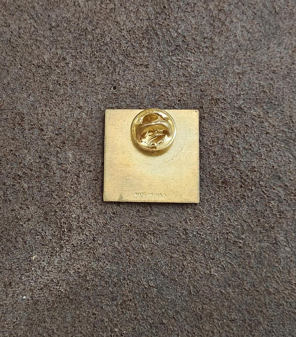 Back of a square gold-tone pin with round clasp and small maker text