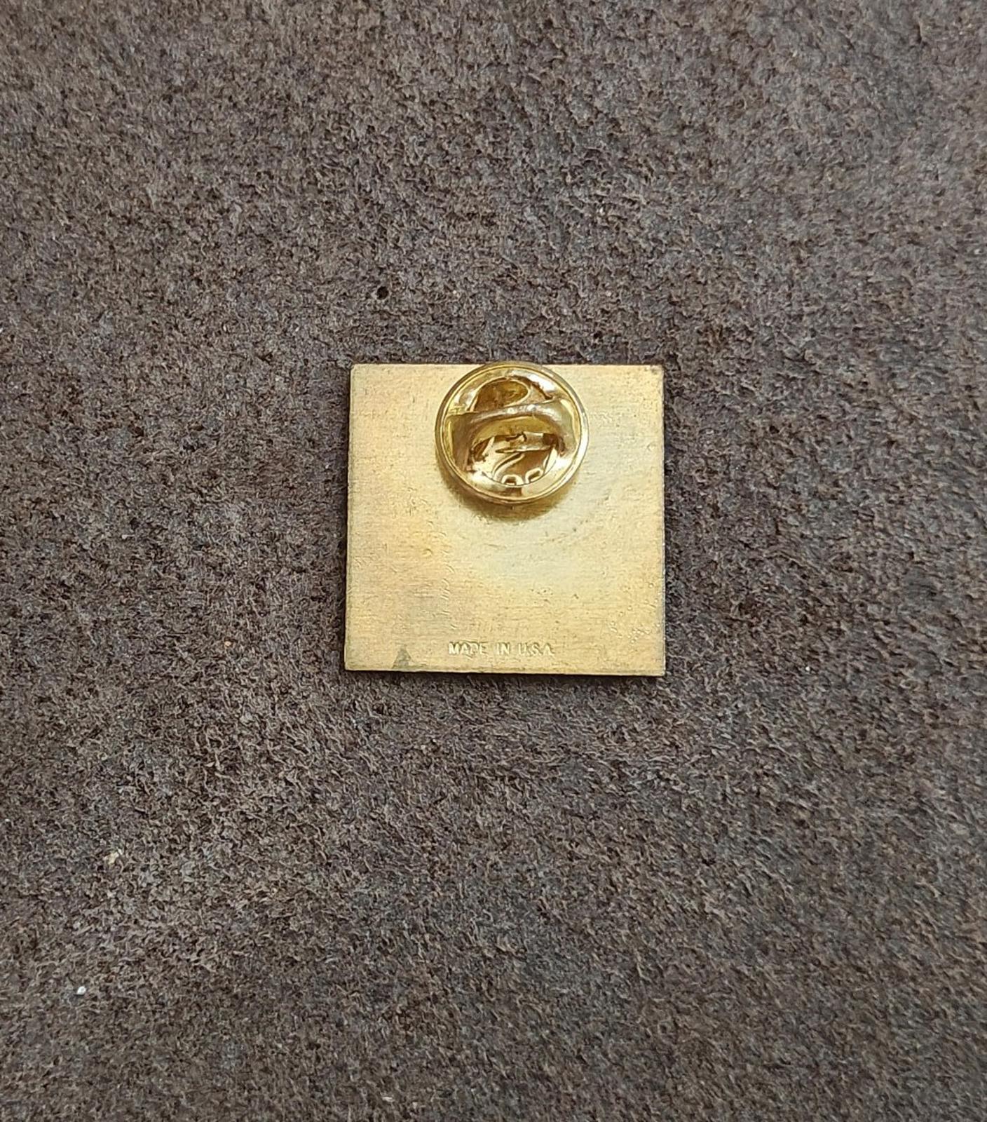 Back of a square gold-tone pin with round clasp and small maker text