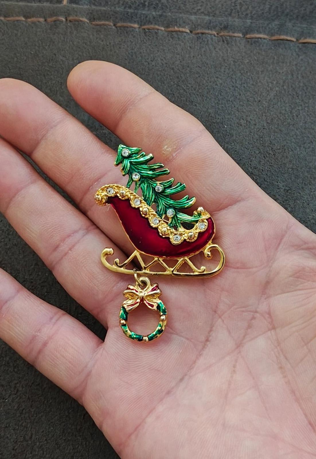 Red sleigh enamel pin with green tree, gold runners, and hanging wreath dangle on hand.