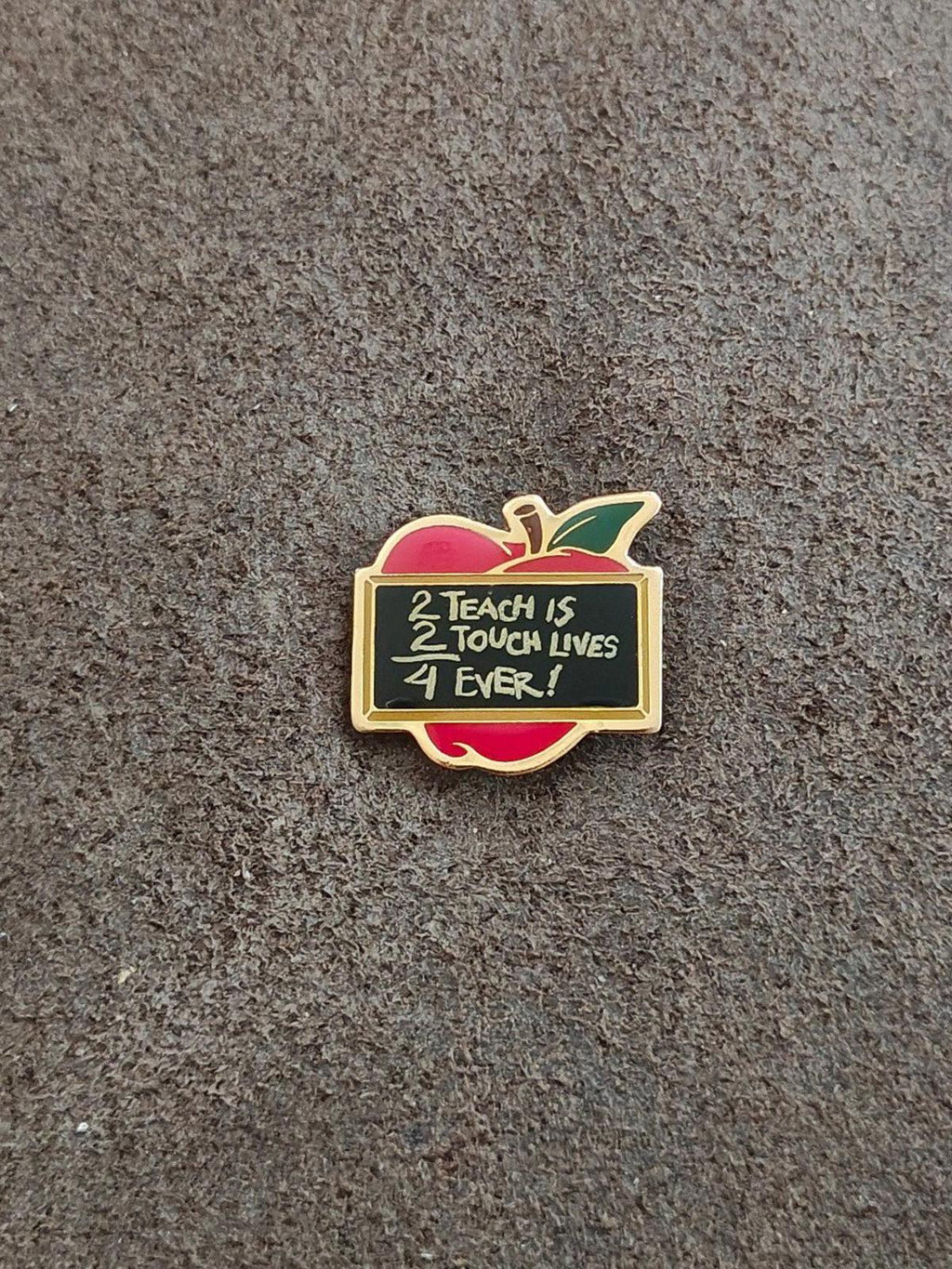 Gold-tone apple teacher pin with red and green enamel and black quote panel on gray background
