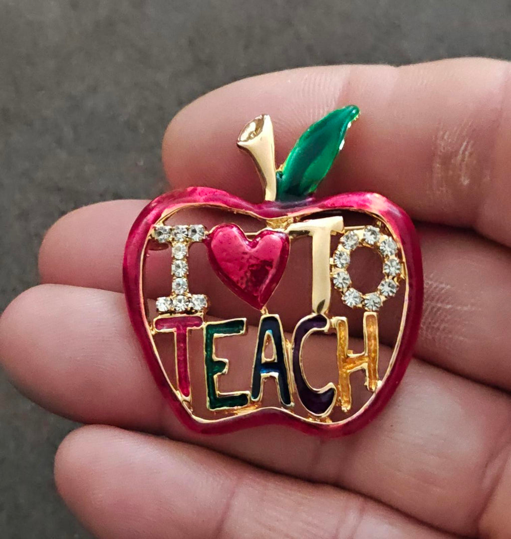 Red enamel apple-shaped brooch with gold tone frame, rhinestone accents, "TEACH" text, heart.
