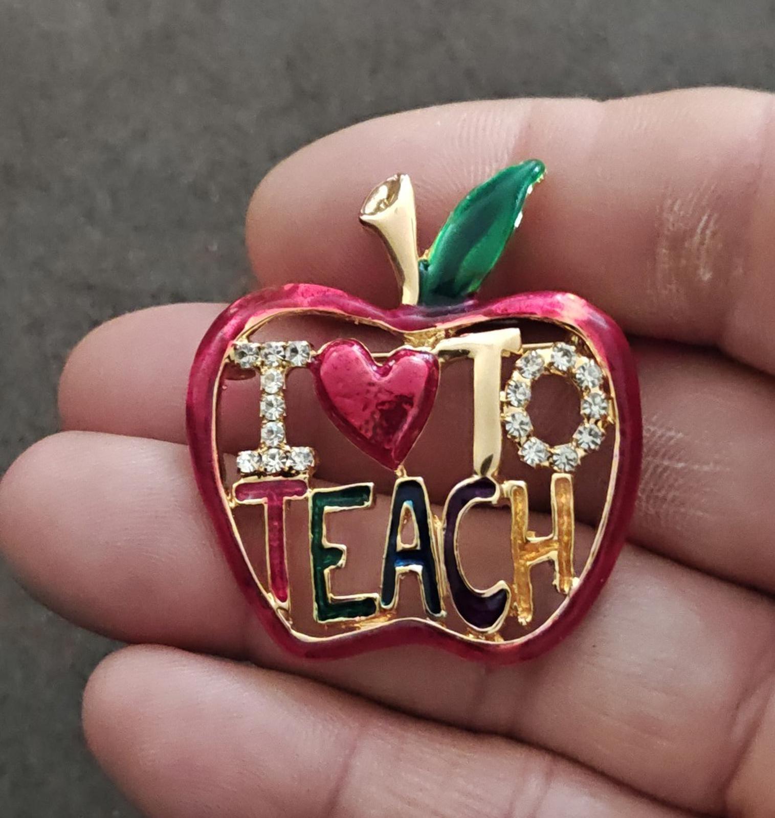 Red enamel apple-shaped brooch with gold tone frame, rhinestone accents, "TEACH" text, heart.