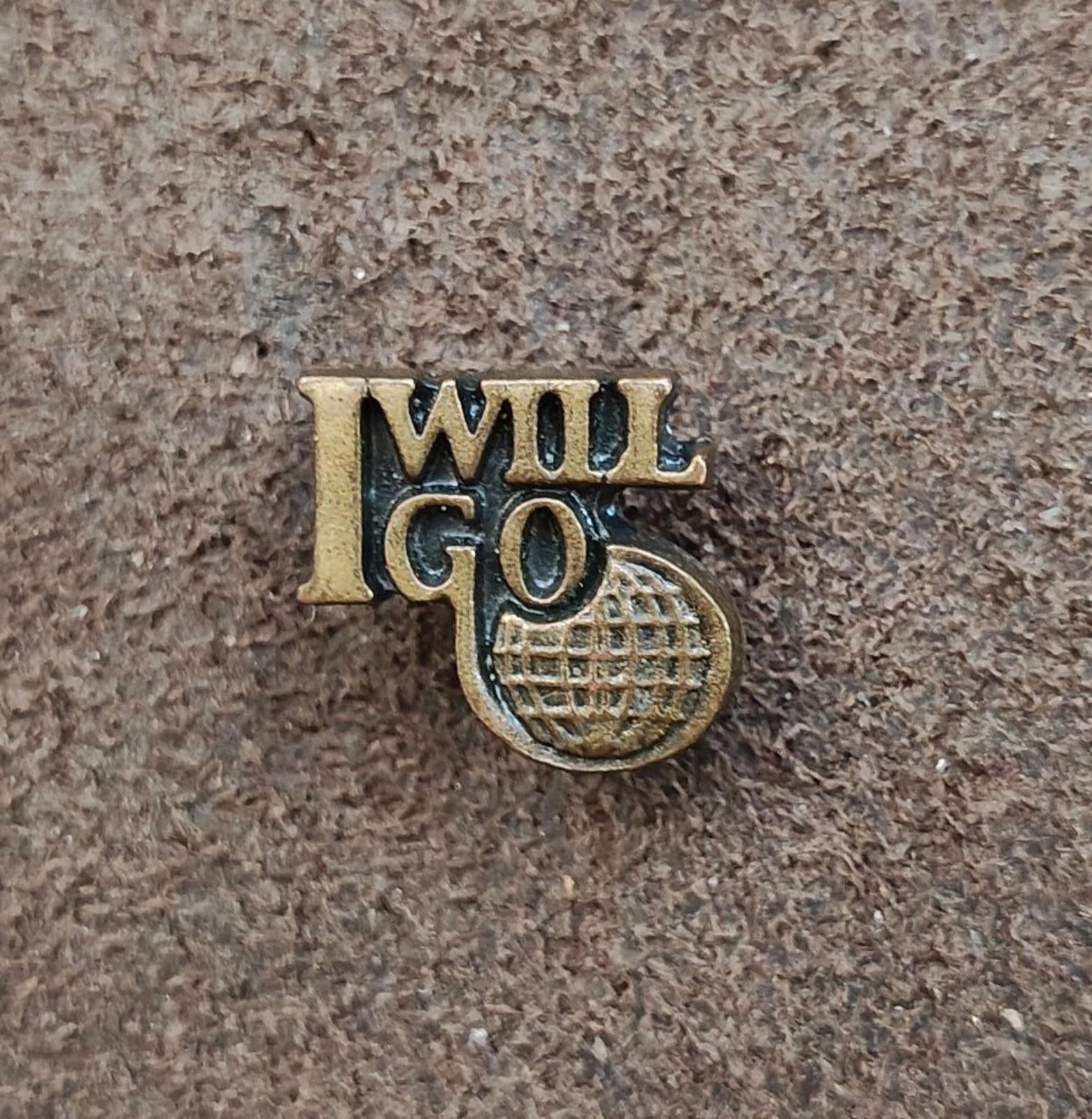 Gold tone "I WILL GO" lapel pin with globe-style emblem, front view on textured surface