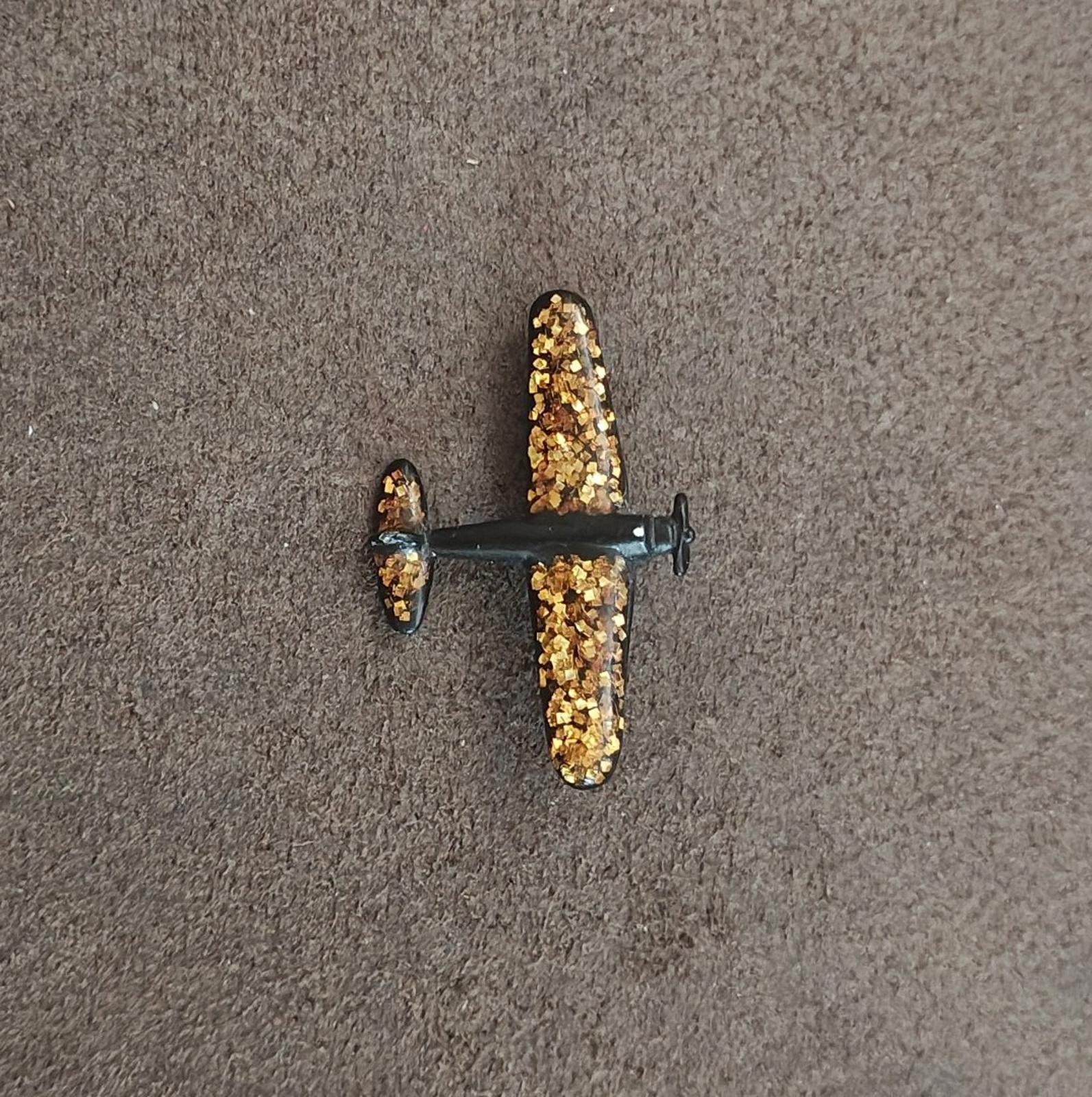 Gold glitter airplane brooch with dark frame, shown flat on dark fabric