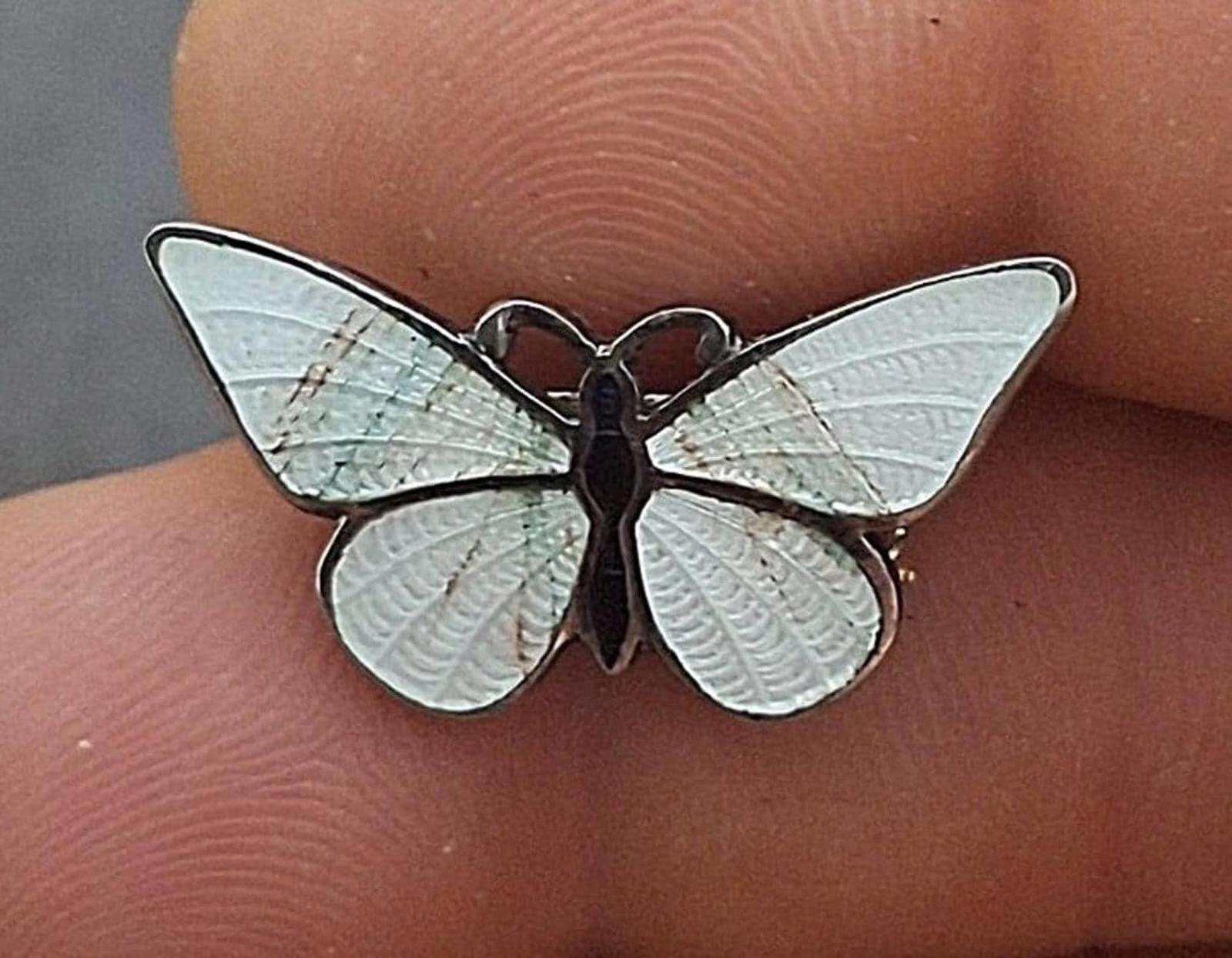 Enamel butterfly pin brooch with white wings and dark body, worn on fingertip.