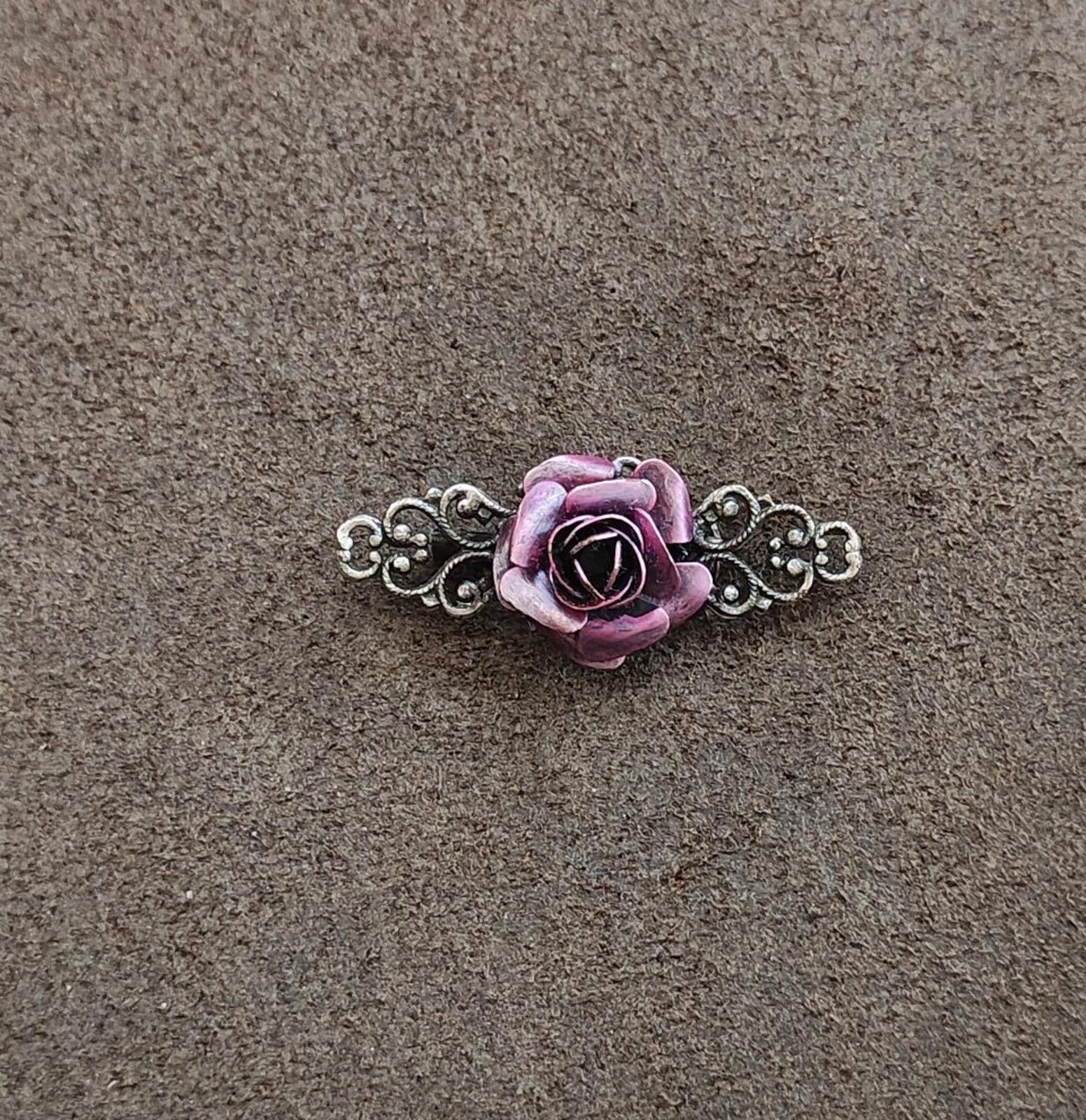 Purple rose brooch with silver tone scrollwork bar, laid on a textured gray surface
