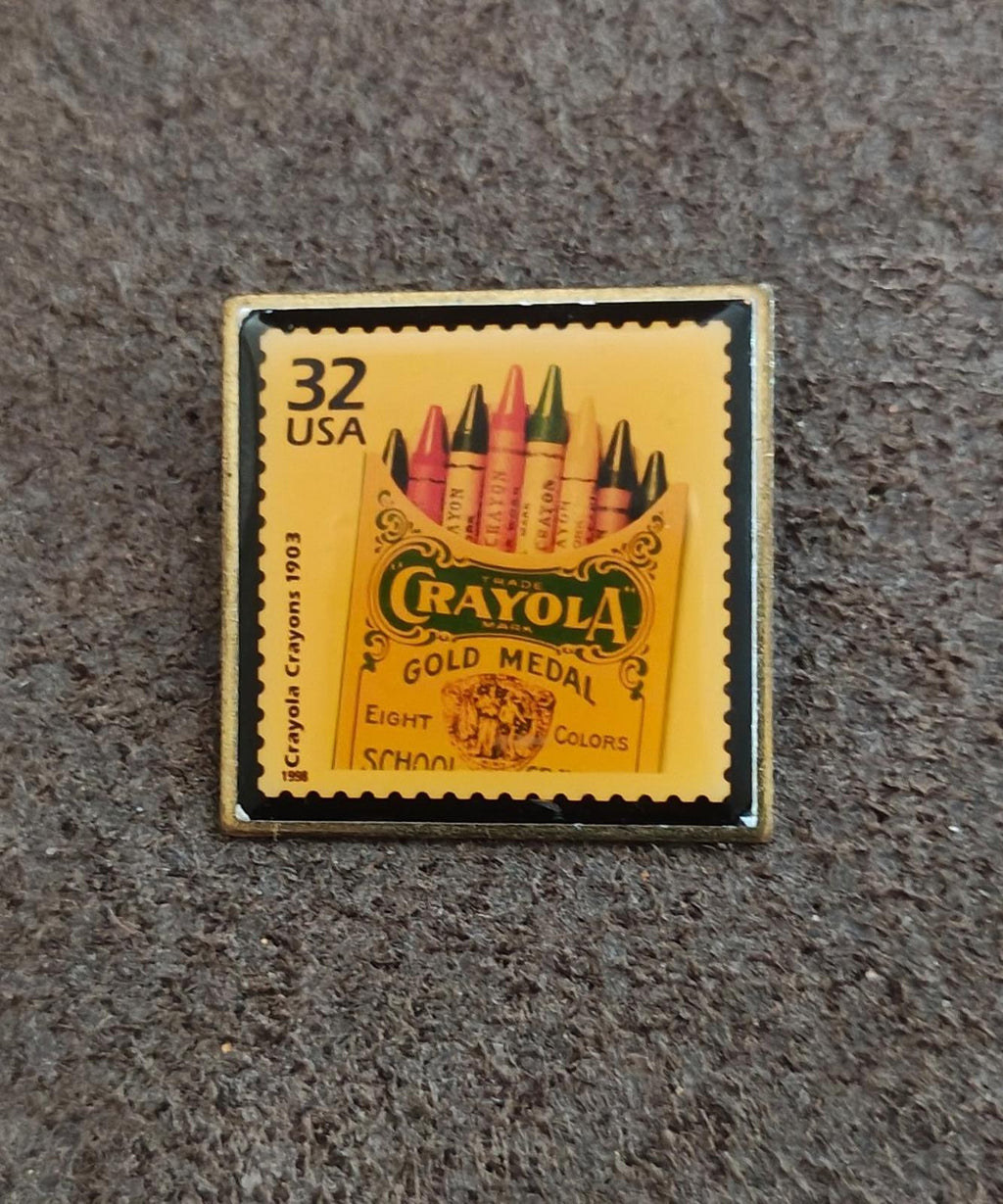 Square gold-tone pin with yellow USPS-style stamp reading “Crayola Gold Medal”