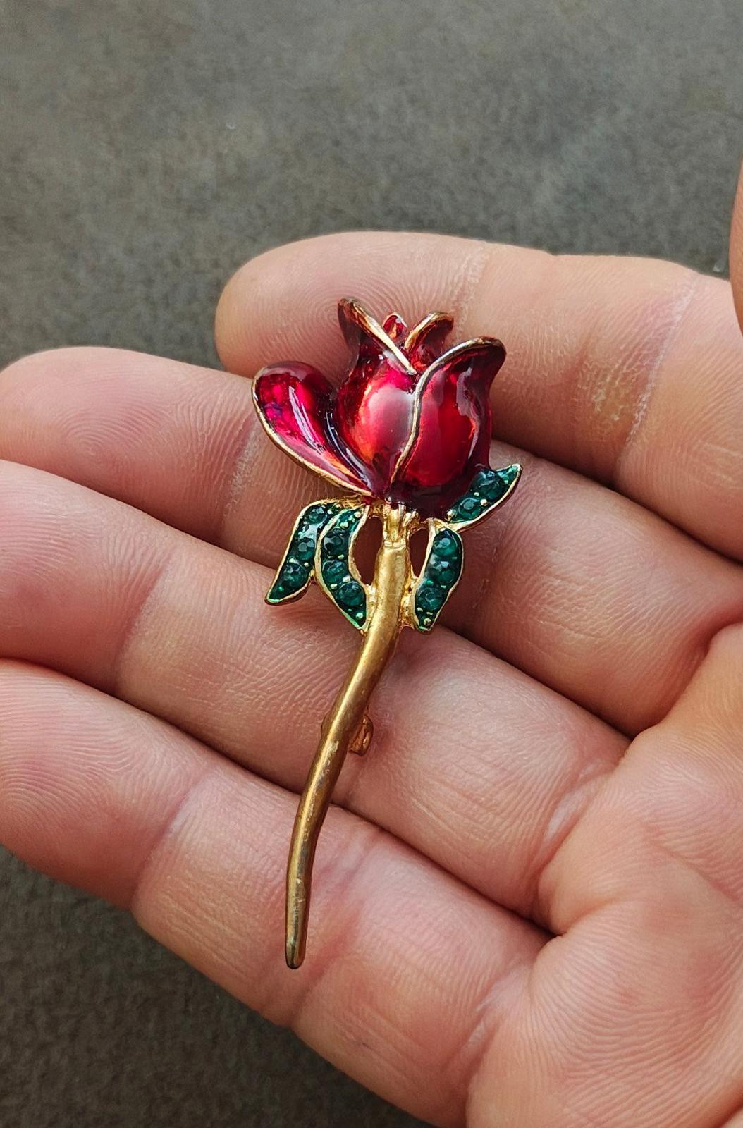 Red enamel flower brooch with green rhinestone leaves and gold-tone stem, close on fingers