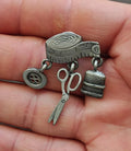 Pewter sewing-theme dangle brooch held in hand, featuring spool, button, scissors, and thimble
