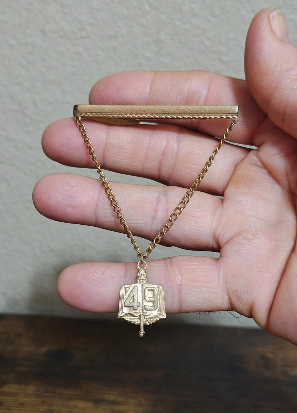 Gold-tone tie clip with chain and book charm marked 49, held in hand