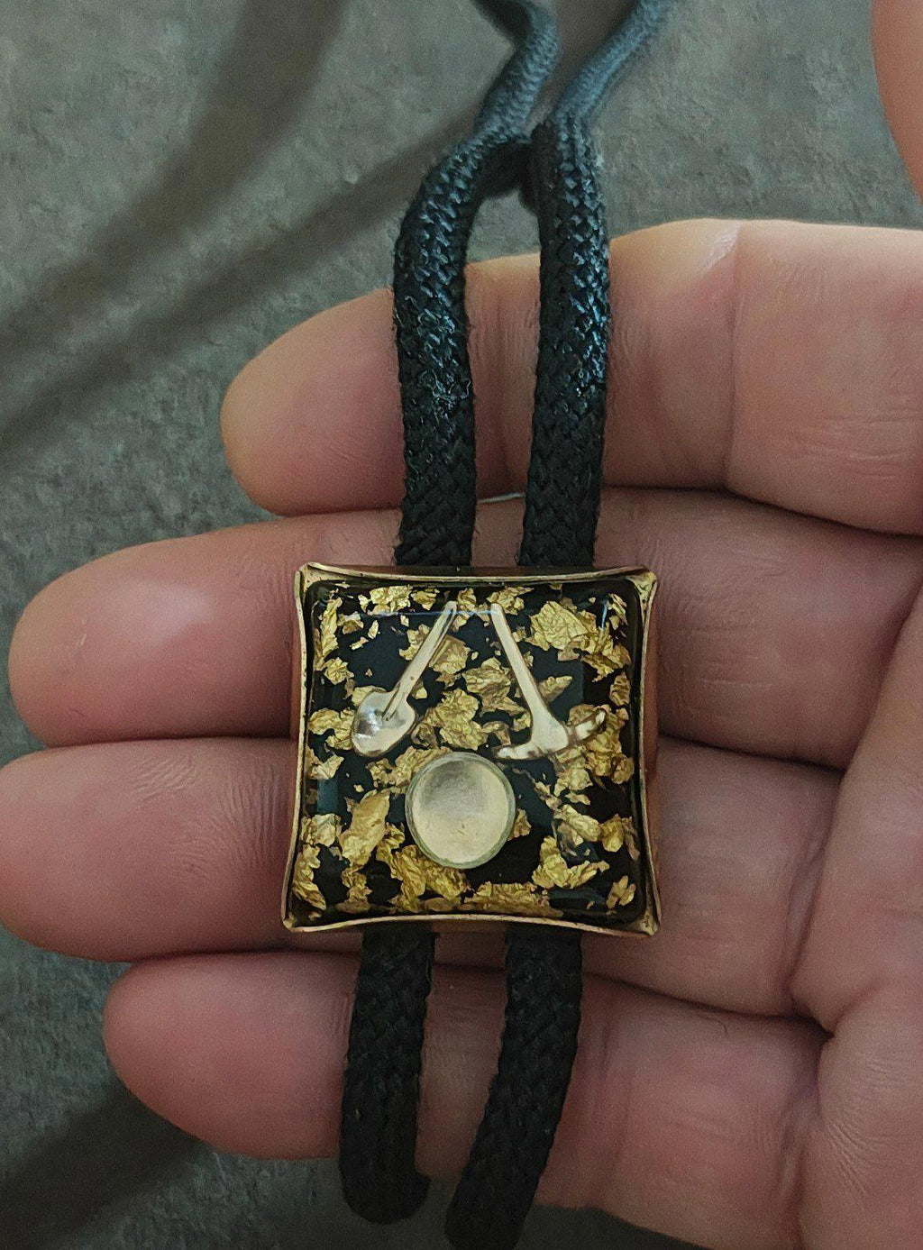 Gold square bolo pendant with gold flecks and round disc, held over black cord