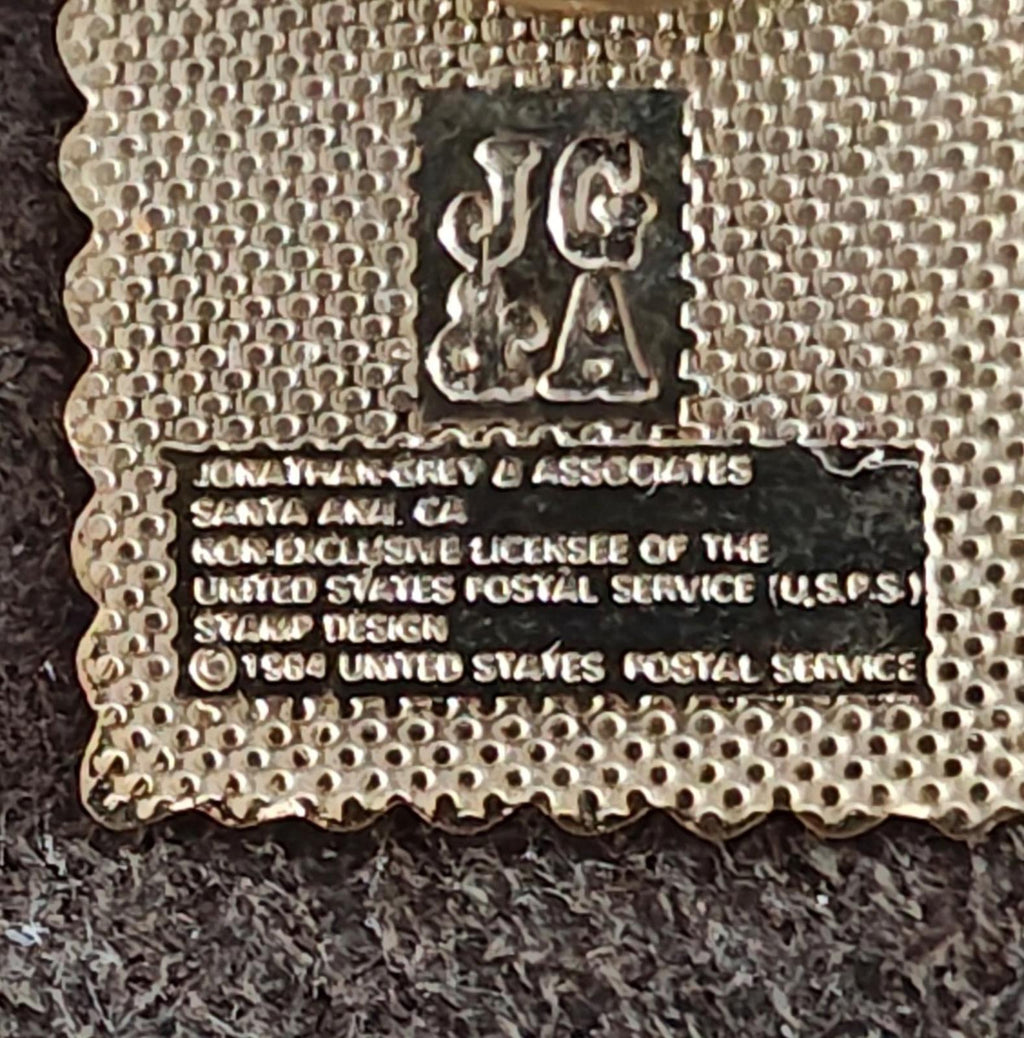 Gold-tone pin back with stamped text and logo on textured metal surface.