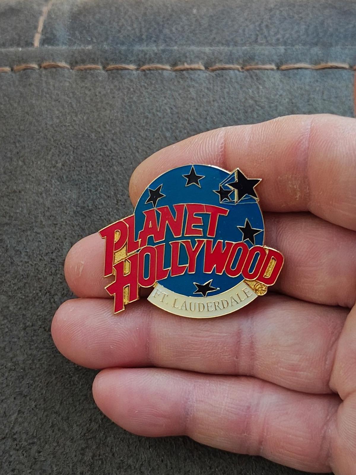 Blue enamel lapel pin with red Planet Hollywood text and stars, gold rim, held in hand.