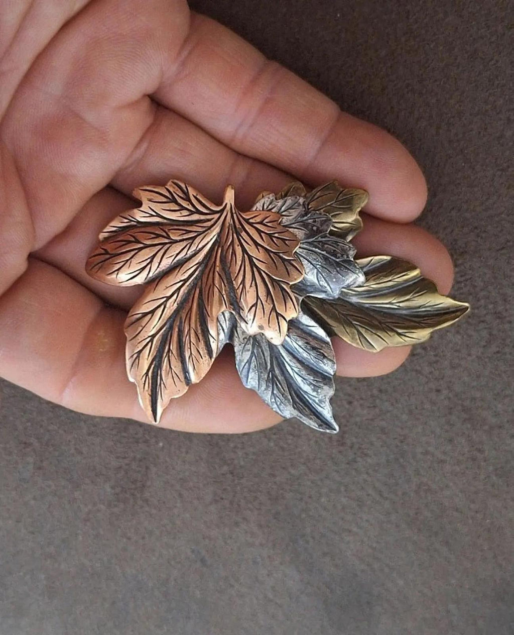 Layered leaf pin brooch with brown, gold, and silver leaves held in an open palm