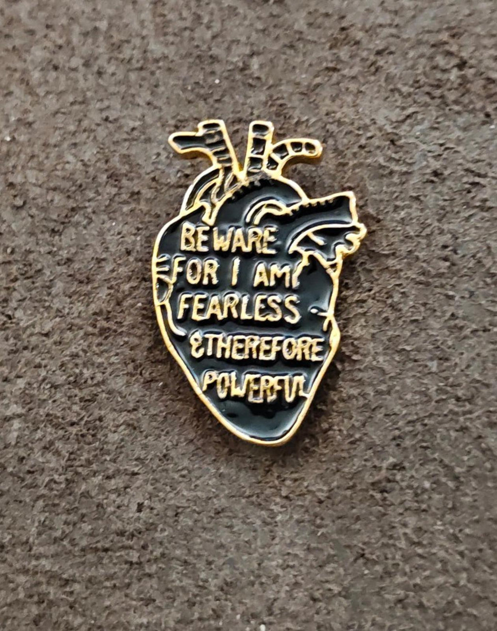 Gold-tone anatomical heart enamel pin with black enamel warning text, centered on gray