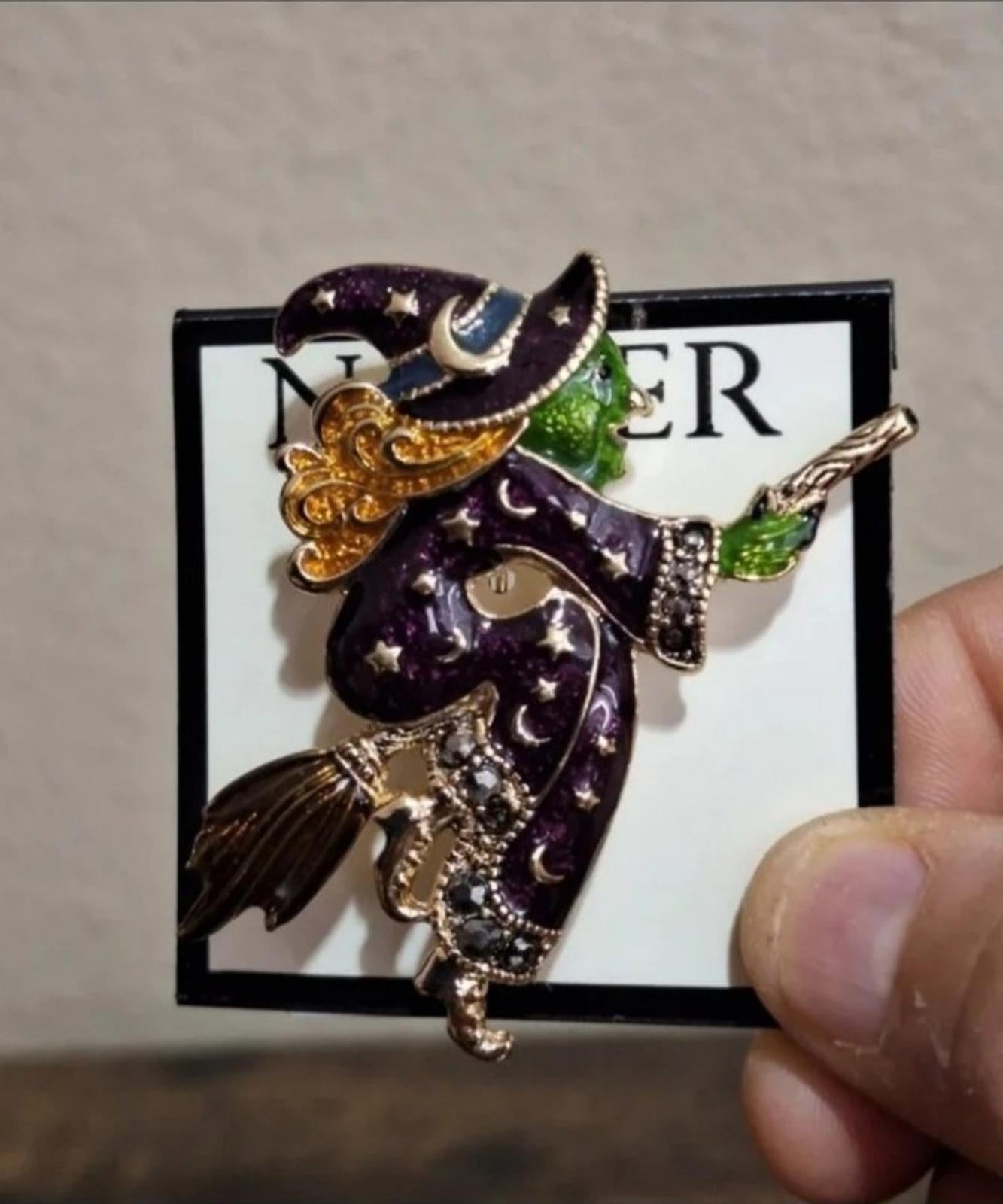 Gold-tone witch enamel pin with dark purple cloak, green accents, and broom
