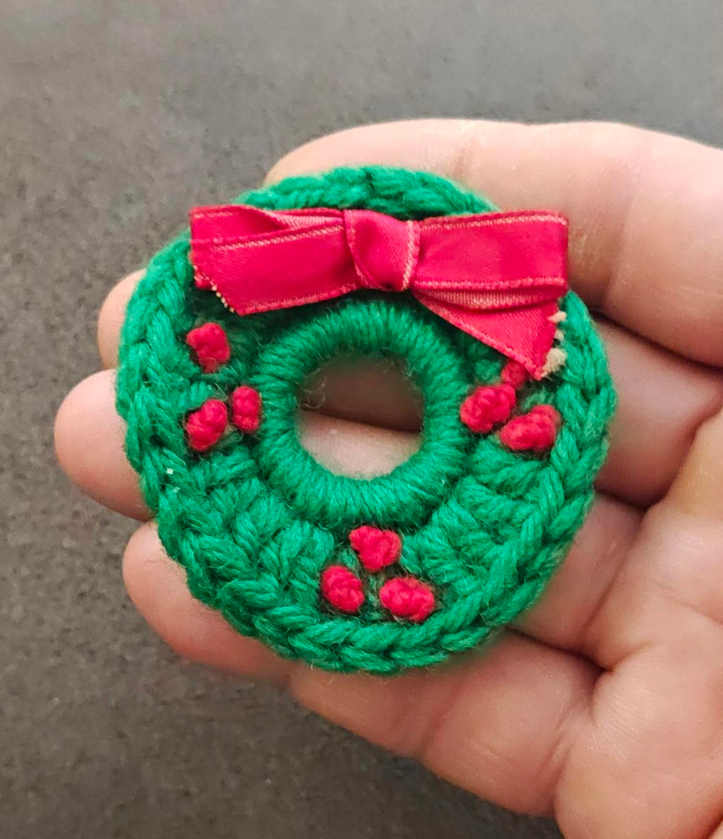 Green knitted wreath brooch with pink bow, red berry accents, round ring front close-up