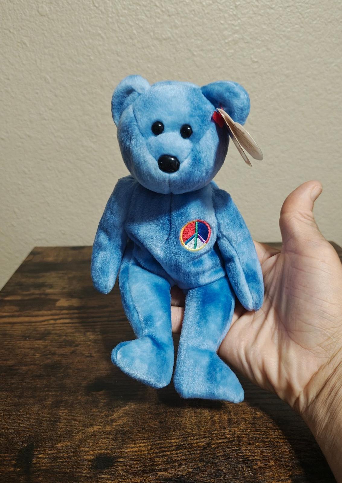 Blue bear plush with peace symbol patch, beanbag style, holding a red ribbon tag