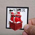 Red enamel Santa climbing down chimney pin on branded card, held by fingertip