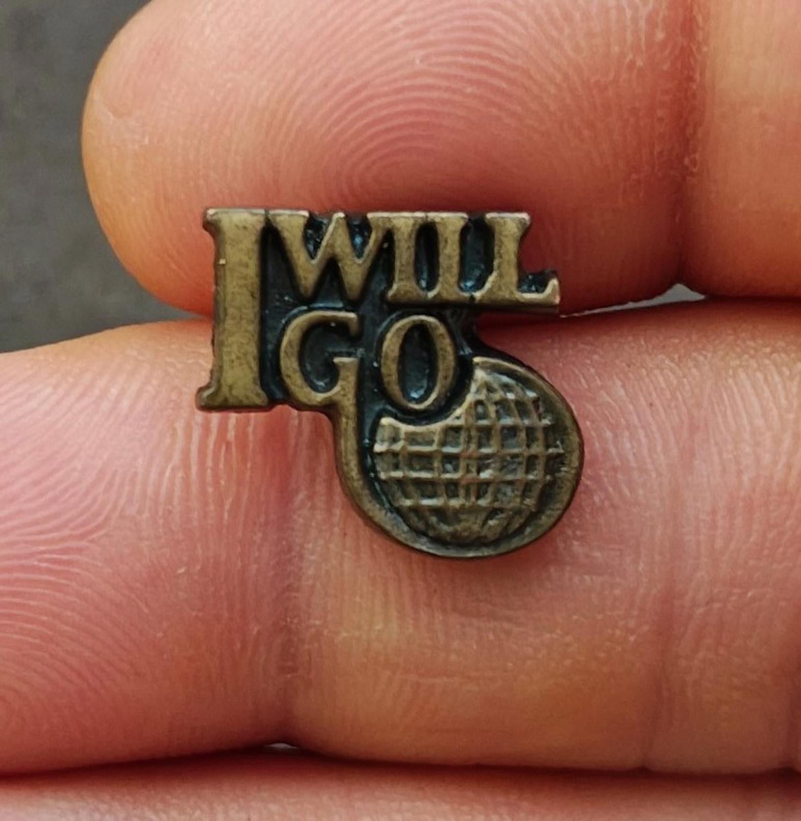 Gold tone lapel pin with "I WILL GO" text and globe-style emblem, front close-up